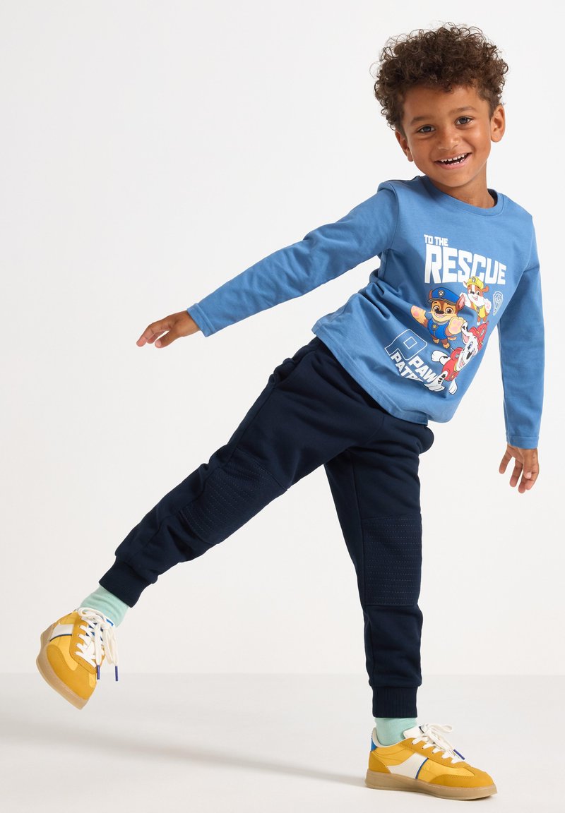 Blaues Langarmshirt mit einem Cartoon-Graphic, kombiniert mit dunkelblauen Jogginghosen mit strukturierten Einsätzen. Gelbe Sneakers mit weißen Akzenten.