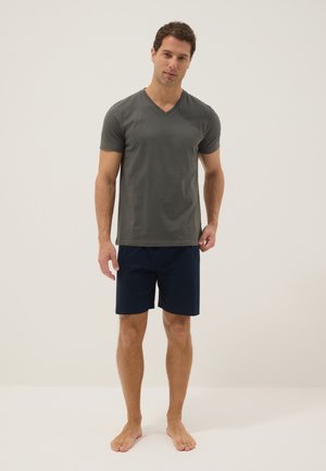 Hombre de pie descalzo, vistiendo una camiseta de manga corta de color gris oscuro con cuello en V y pantalones cortos azul marino sobre un fondo claro y liso.