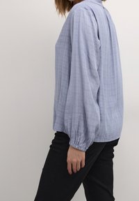 Chemise bleu clair à boutons avec un tissage texturé, manches bouffantes et poignets froncés, associée à un pantalon noir.