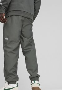 Szare sportowe spodnie z elastycznymi mankietami, z odblaskowymi naszywkami, gładką fakturą i luźnym krojem. Stylizowane z białymi trampkami.