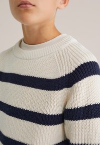 Sweter dziany w kolorze kremowym z granatowymi poziomymi paskami. Posiada prążkowany okrągły dekolt oraz teksturowany materiał. Zbliżenie na ramiona.