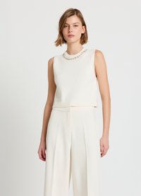 Ärmelloser weißer, kurz geschnittener Pullover mit geripptem Saum und mit Perlen besetztem Ausschnitt, kombiniert mit cremefarbenen, weit geschnittenen Hosen. Glatte Stricktextur.