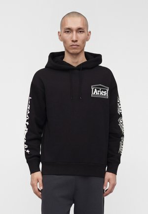 Férfi, fekete kapucnis pulóverben, fehér "Aries" logóval a mellkason és fehér grafikákkal mindkét ujján, sima háttér előtt áll.