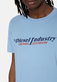 Jasnoniebieska bawełniana koszulka z dużym nadrukiem z napisem „Diesel Industry” w granatowym kolorze oraz „DENIM DIVISION” w czerwonym. Klasyczny okrągły dekolt.