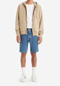Beige zip-up hoodie med en framficka, bärs över en vit t-shirt, ihop med blå denimshorts och vita strumpor, avslutat med vita sneakers.