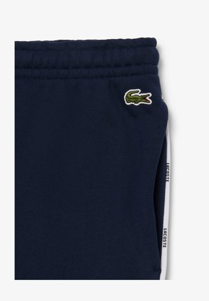 Tumed sinised trenningpüksid elastse vöökohtadega, millel on roheline tikandiga krokodilli logo ja valged külje triibud, millel on tekst "LACOSTE".