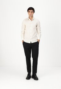 Camasă bej deschis cu guler și un model subtil, asortată cu pantaloni negri și pantofi negri cu șireturi. Modelul stă cu mâinile în buzunare.