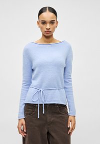 Pull bleu clair au tissu doux avec un large col bateau et des manches longues, présentant un cordon de serrage à la taille. Associé à un pantalon foncé.