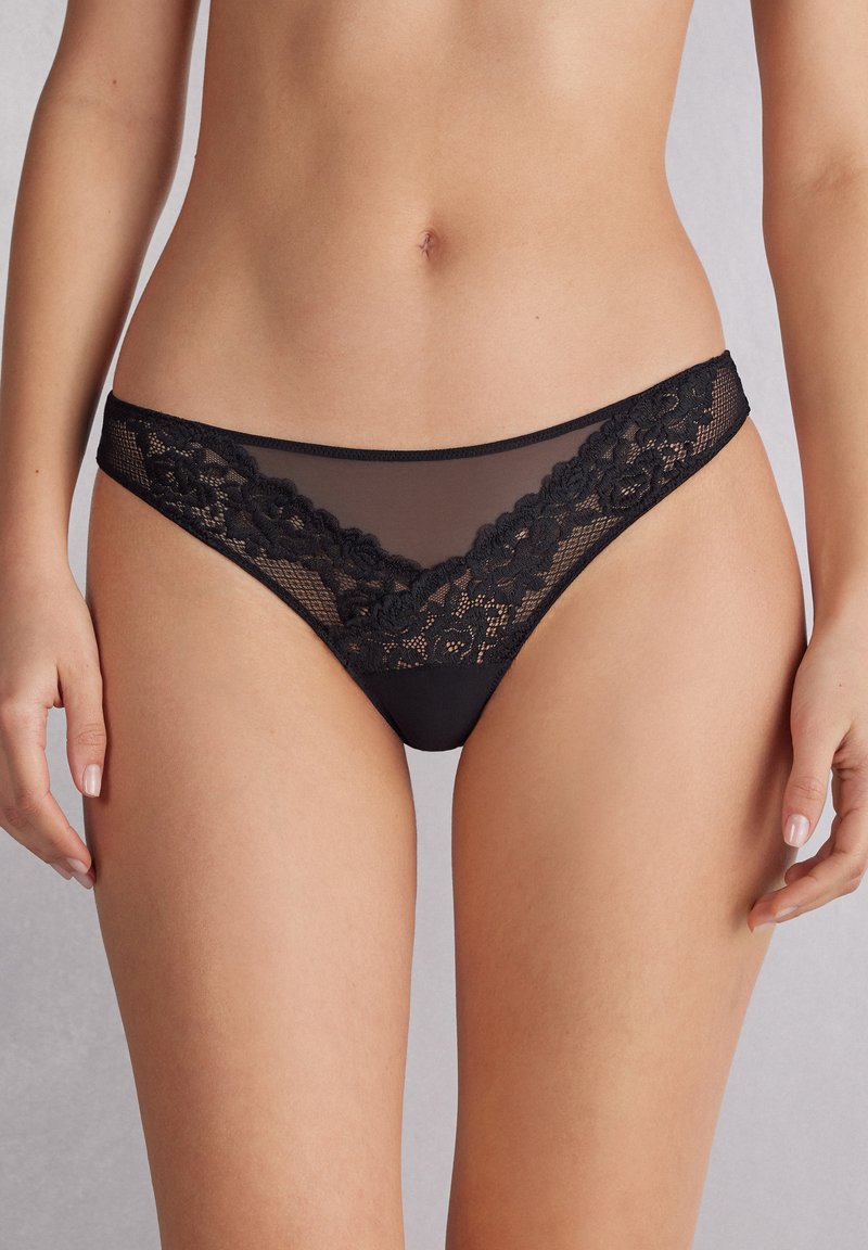 Svart spetsstringtrosor med en genomskinlig meshpanel med blommönster, släta kanter och ett kontrasterande midjeband.
