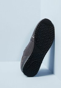 En mörkgrå sneaker lutad mot en ljusblå vägg, som visar den detaljerade svarta gummisolen med texturerade greppmönster.