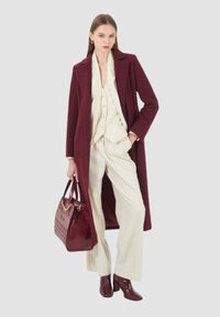 Cappotto di lana bordeaux su un completo crema, con un gilet a tre bottoni, pantaloni larghi, borsa coordinata e stivali lucidi fino al ginocchio.