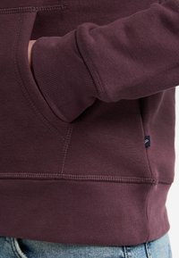 Bordeauxfarbener Hoodie aus weichem Baumwollgemisch, mit gerippten Bündchen und einer Kängurutasche. Kleinere Logodetails am Saum. Legere Passform.