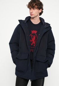 Sötétkék steppelt dzseki kapucnival, cipzáras záródással és két első zsebbel. Az anyag simának tűnik enyhén fényes felülettel.