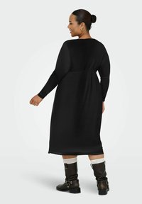 Robe noire à manches longues avec une coupe décontractée, arrivant au genou, et une taille froncée. Associée à des bottes noires jusqu'aux genoux et à des chaussettes blanches texturées.