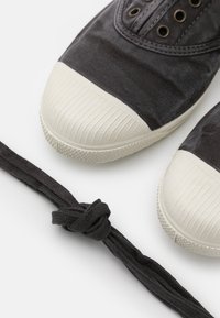 Baskets en toile noires avec avant en caoutchouc blanc et semelle texturée ; inclut un lacet noir noué, mettant en avant des caractéristiques de design simple.