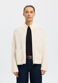 Cárdigan de lana beige claro con cuello chal, dos bolsillos delanteros y puños acanalados, con un ajuste relajado y una textura suave.