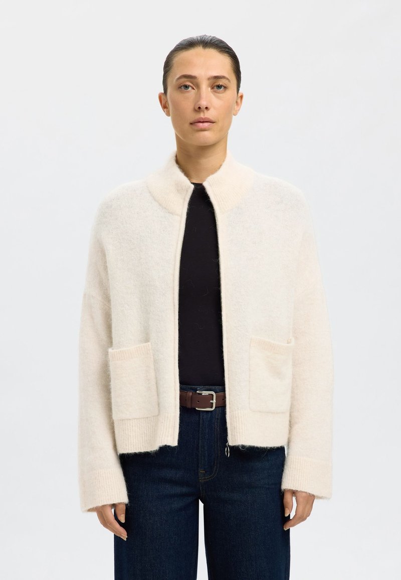 Cárdigan de lana beige claro con cuello chal, dos bolsillos delanteros y puños acanalados, con un ajuste relajado y una textura suave.