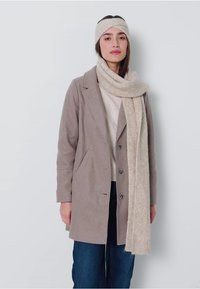Manteau en laine beige avec un col cranté, fermeture à boutons, associé à une écharpe beige claire. Le modèle porte un bandeau assorti, se tenant devant un fond uni.