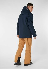 Giacca impermeabile blu navy con cappuccio, abbinata a pantaloni da neve color sabbia con accenti neri. Tessuto liscio, vestibilità su misura e design pratico.