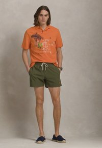 Camisa polo laranja com estampado gráfico, combinada com calções verdes-azeitona. Inclui cintura elástica com cordão; modelo usa espadrilhas azul-marinho.