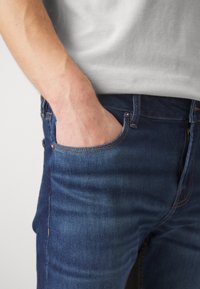 Sötétkék farmernadrág, testhez álló fazonnal, látható varrásokkal, és egy kéz pihen a hátsó zsebben.