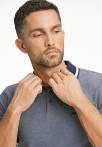 Polo grigio con colletto blu navy, tessuto strutturato e una placca con tre bottoni. Ha un aspetto leggermente aderente, con accenti scuri sul logo.