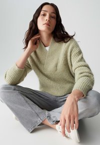 Helles, grün strukturiertes Strickoberteil mit V-Ausschnitt und gerippten Ärmeln, kombiniert mit grauen weit geschnittenen Hosen und weißen Sneakers.