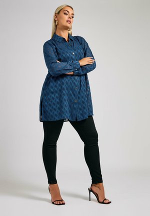 Blue patterned long shirt made of a lightweight material, button-up design with a curved hem, paired with black skinny pants and strappy heels.
