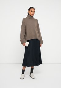 Femme portant un pull oversize taupe à col roulé, une jupe midi plissée noire, des chaussettes grises et des mocassins blancs, tenant une petite pochette tissée blanche.