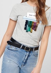 Šedé tričko s krátkým rukávem zobrazuje grafiku se flitry s textem "ŠPATNÉ VIBRACE" a barevné svislé pruhy. Nosené s černým opaskem.