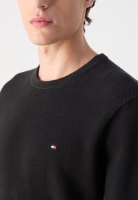 Zwarte gebreide trui met een ronde halslijn, voorzien van een klein rood, wit en blauw logo op de borst, met een gestructureerde stof.