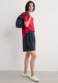Rotes T-Shirt mit kurzen Ärmeln, kombiniert mit dunkelblauen, gefältelten Shorts. Weiße Sneakers vervollständigen das Outfit, zusammen mit einem dunkelblauen Rucksack, der über die Schulter gehängt ist.