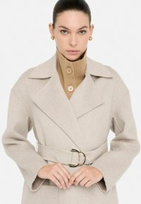 Cappotto in misto lana beige con ampi revers, vita con cintura e tessuto testurizzato. Abbinato a un dolcevita a coste color beige con dettagli a bottone.