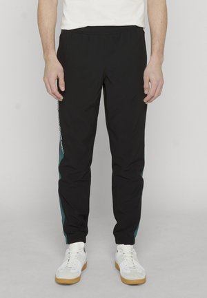 Mann iført svarte joggebukser med turkise sidepaneler og hvite joggesko, stående mot en lys, ensfarget bakgrunn.