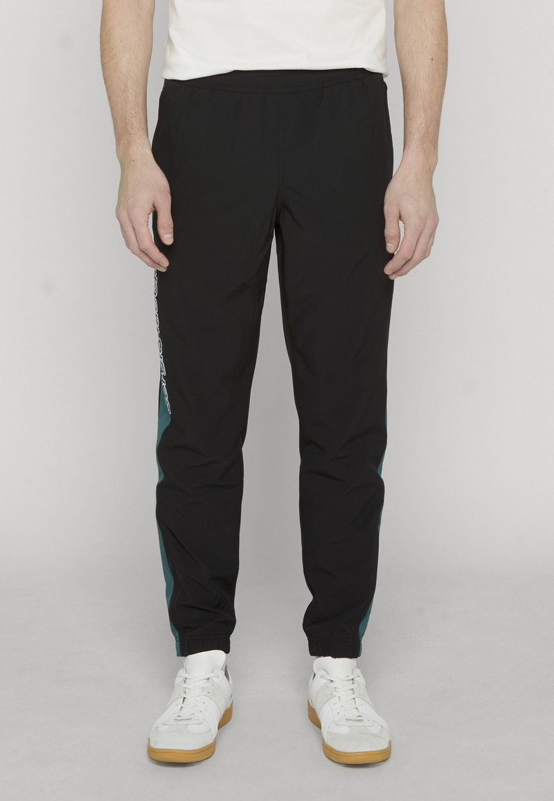 Man draagt zwarte joggerbroek met turkooize zijpanelen en witte sneakers, staand tegen een effen lichte achtergrond.