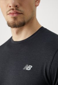 Fekete sportpóló texturált mintával, kisebb, fehér New Balance logóval a mellrészen. Kerek nyakú és testhezálló szabású.