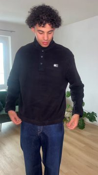 Νέος άνδρας με σγουρά μαλλιά που ρυθμίζει ένα μαύρο πουκάμισο με κουμπιά, στέκεται σε ένα φωτεινό δωμάτιο με ξύλινο δάπεδο και πράσινα φυτά.