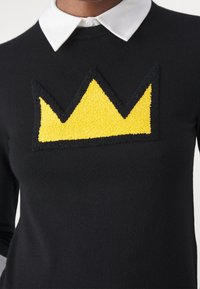 Pull noir à manches longues avec un motif texturé de couronne jaune sur la poitrine, accentué par un col blanc.