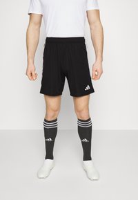 Zwarte sportshorts met een elastische tailleband, voorzien van witte Adidas-logo. Gecombineerd met knieën-hoge zwarte sokken met witte strepen.