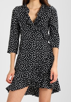 Femme portant une robe portefeuille noire à pois blancs irréguliers, manches trois-quarts, col à volants et ourlet asymétrique.