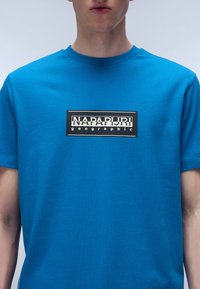 Kortærmet blå t-shirt lavet af bomuld, med et rektangulært sort logo med hvid tekst centreret på brystet. Glat tekstur.