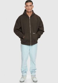 Haut à capuche zipé marron avec de grandes poches et un cordon de serrage, associé à un jean bleu clair et des baskets blanches. Texture lisse et coupe décontractée.