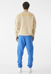 Beige genser med oransje tekst på baksiden, kombinert med blå joggebukser. Modellen har på seg hvite sneakers. Glatt tekstur gjennom hele.