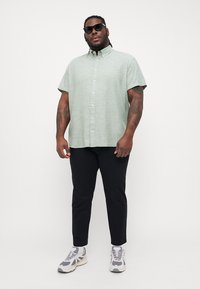 Cămașă scurtă, cu mâneci scurte, de culoare verde deschis, cu guler, purtată cu pantaloni negri și pantofi sport gri. Cămașa are o textură subtilă și nasturi.
