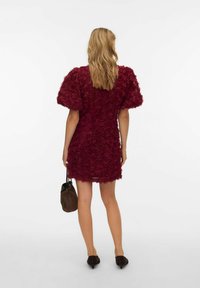 Bordowa sukienka z trójwymiarowymi aplikacjami róż, bufkami na rękawach, dopasowaną sylwetką i przezroczystym materiałem wykończeniowym. W zestawieniu z brązową torebką.