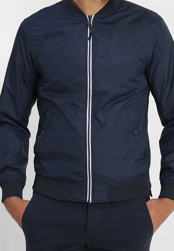 Homme portant une veste bomber bleu foncé avec une fermeture éclair à l'avant, des poches latérales et des poignets côtelés, debout les mains dans les poches.