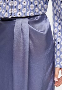 Ein schimmernder Lavendelrock mit glatter Textur und einem Frontknoten-Detail, kombiniert mit einer gemusterten Bluse in Blau und Weiß mit floralen Motiven.