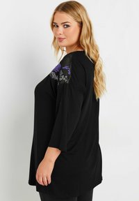 Schwarzes Tunika-Oberteil mit drei Viertel-Ärmeln, das blumende Stickerei in Lila und Weiß an den Schultern aufweist. Weicher, dehnbarer Stoff.