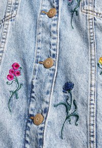 Jeanskappa i ljusblått tyg med blommig broderi i rosa, rött, gult och blått, samt ljusbruna, vävda knappar.