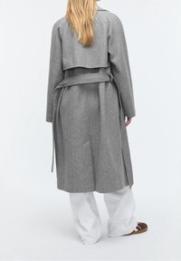 Personne aux longs cheveux blonds portant un manteau en laine gris ceinturé, un pantalon large blanc et des chaussures marron, debout de dos sur un fond blanc.